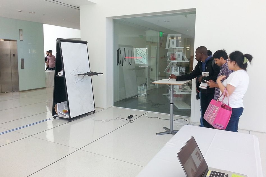 Students in the MIT Media Lab try out a pen-controlled "writing" drone. When users trace lines on a desk with an inkless pen, the drone will duplicate those lines on a distant whiteboard. This is done with cameras tracking the pen and drone, and software synching the two patterns.