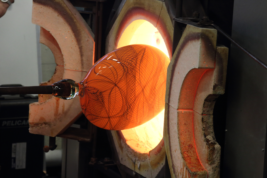 Lollipop look-alike: A glass vessel in progress looks like a giant lollipop as it is pulled out of a glory hole, the furnace for reheating glass while it is being formed. Orange when hot, the glass will cool to a seafoam green with a swirling blue pattern.