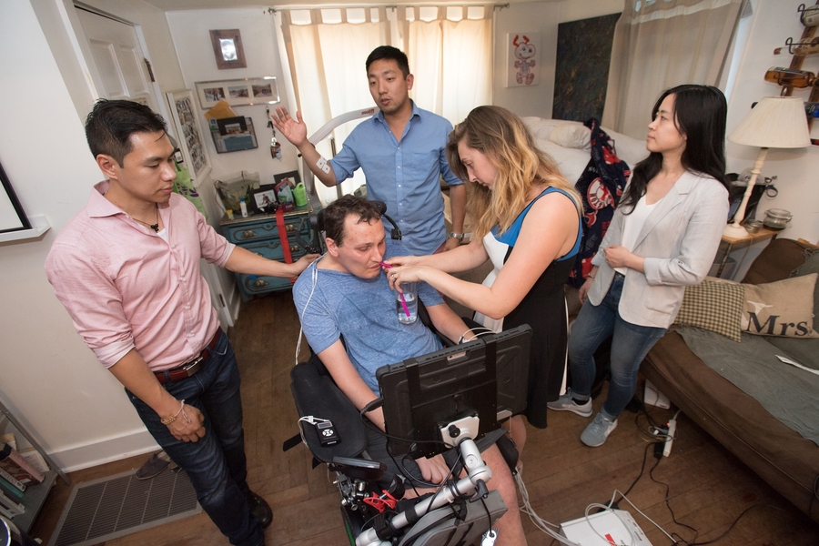 Dexter Ang, left, rests a hand on the back of Bobby Forster, who sips water held by his wife, Casey. Behind them, David Cipoletta, a robotics engineer, tests the sensitivity of the EMG sensors. Wenxin Feng looks on.