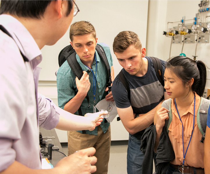 Associate Professor Juejun (JJ) Hu shows a metasurface lens made with chalcogenide alloys to Summer Scholars Simon Egner, Michael Molinski and Sabrina Shen during a lab tour.