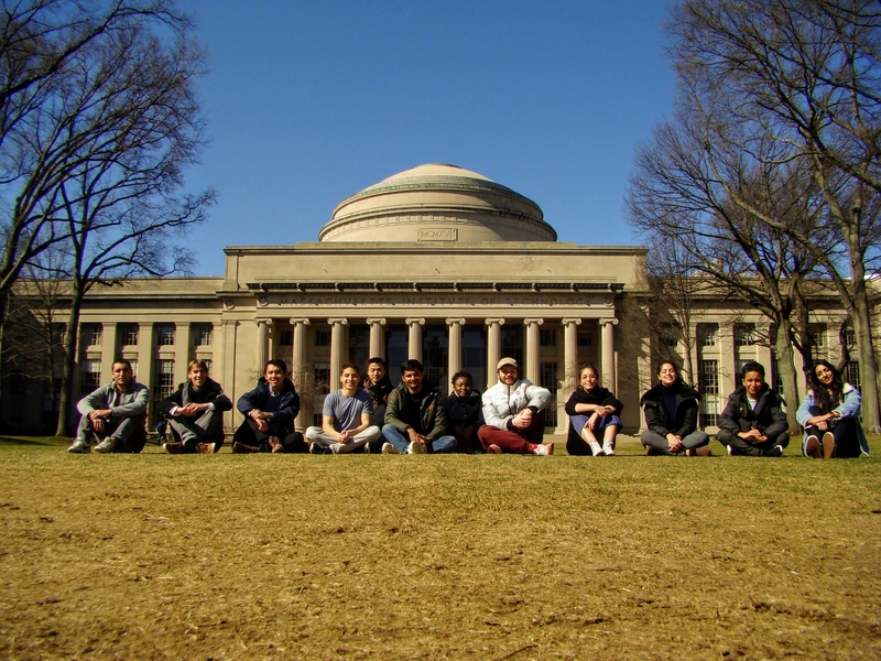 Twelve students sit cross-legged on a lawn in front of MIT's Great Dome during winter