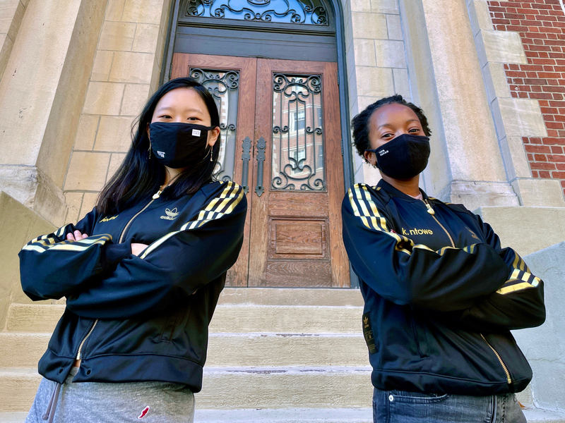 Photo of Jeana Choi and Koumani Ntowe in black sweatshirts and black face masks, posing with arms crossed in front of an MIT building.