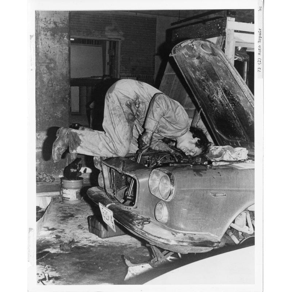 Black and white photo of a student kneeling on an old car, making repairs to the engine