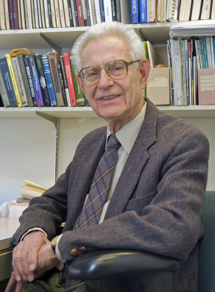 Headshot photo of John Dugundji sitting in an office in front of a bookshelf