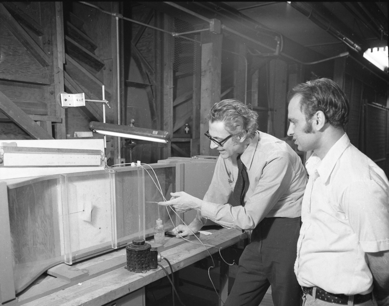 1970s-era photo of Professor Emeritus John Dugundji explaining the impact of vibration on a model of an airplane wing as a graduate student looks on.