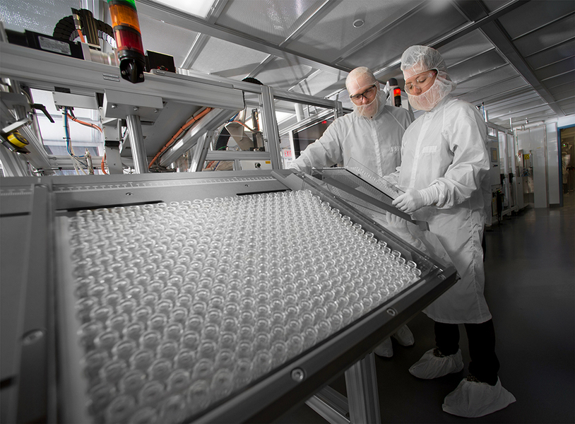 Photo of two technicians covered in white protective coveralls examining a tray of hundreds of little glass vials.