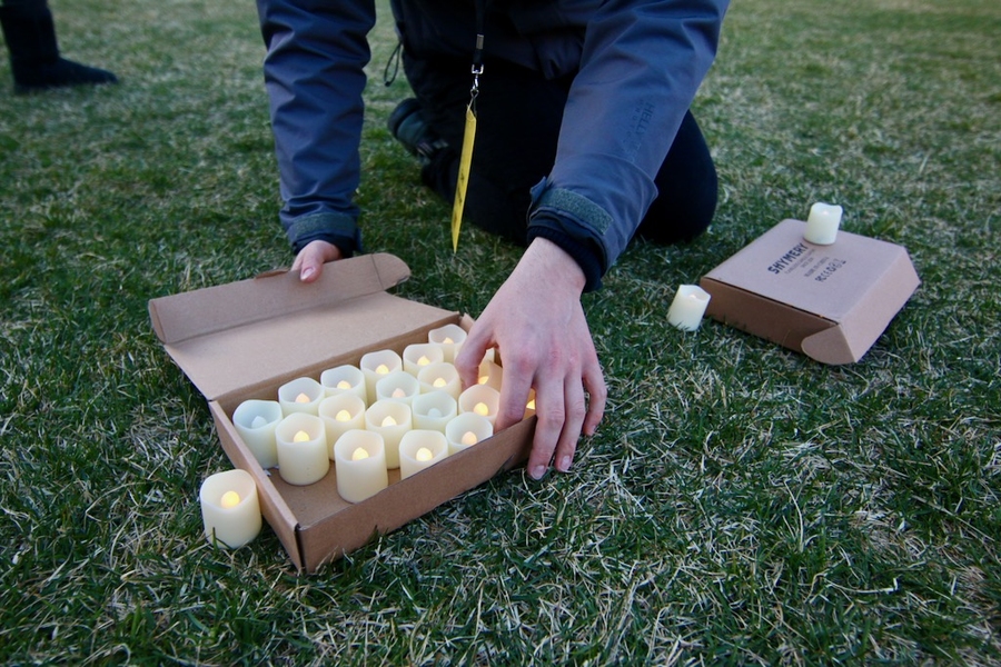 Photo of a box of lit battery-operated candles, with a student volunteer's hand grabbing a candle to place on grass.