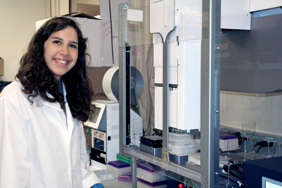 Photo of Mira Moufarrej wearing a lab coat and standing in front of equipment in a lab