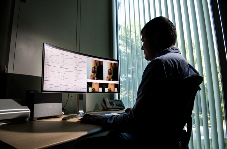 A researcher sits in an office looking at a computer monitor. On the left side of the monitor are several graphs. One the right side are four images of a woman's arm with a crown tattoo.