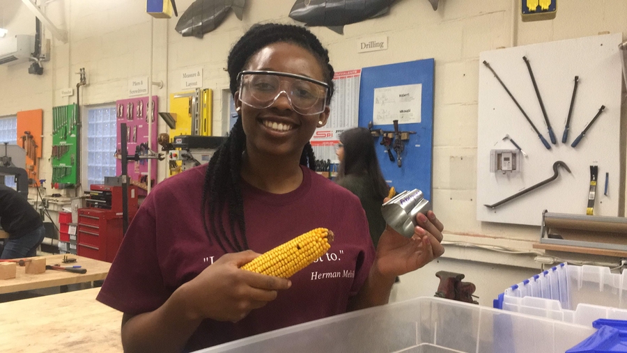 Photo of Abigael Bamgboye, in safety goggles, in a classroom holding an ear of corn in one hand and a metal nozzle in the other.