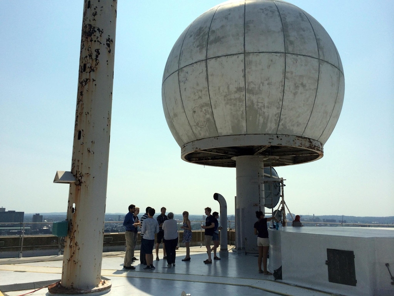 Photo of a large white circular dome pressing against blue sky with a gray pillar in foreground and city in background.