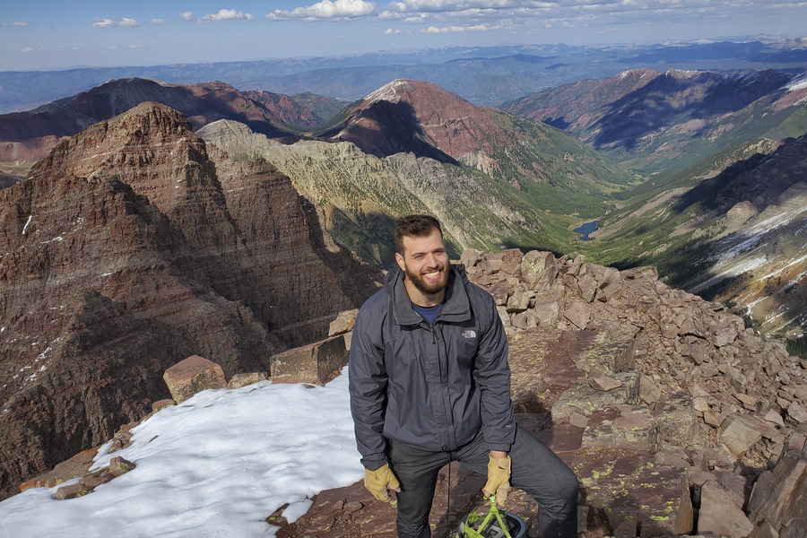 Photo of Theo Mouratidis smiling at the top of a snow-capped mountain, with a vista of mountains and valleys behind him