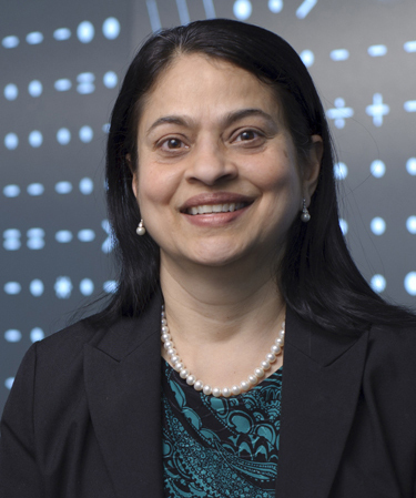 Portrait photo of Anu Agarwal, smiling in front of a dark wall with white mathematical symbols