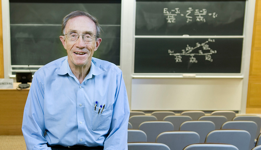 Photo of William Dalzell, smiling and seated before a chalkboard in an empty classroom