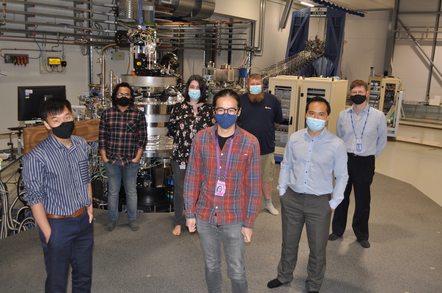 Photo of seven individuals, each wearing facemasks and standing before a large piece of lab equipment.