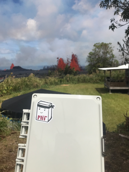 Photo of an air quality sensor, labeled with an MIT sticker, with an active volcanic fissure spewing red lava in the background