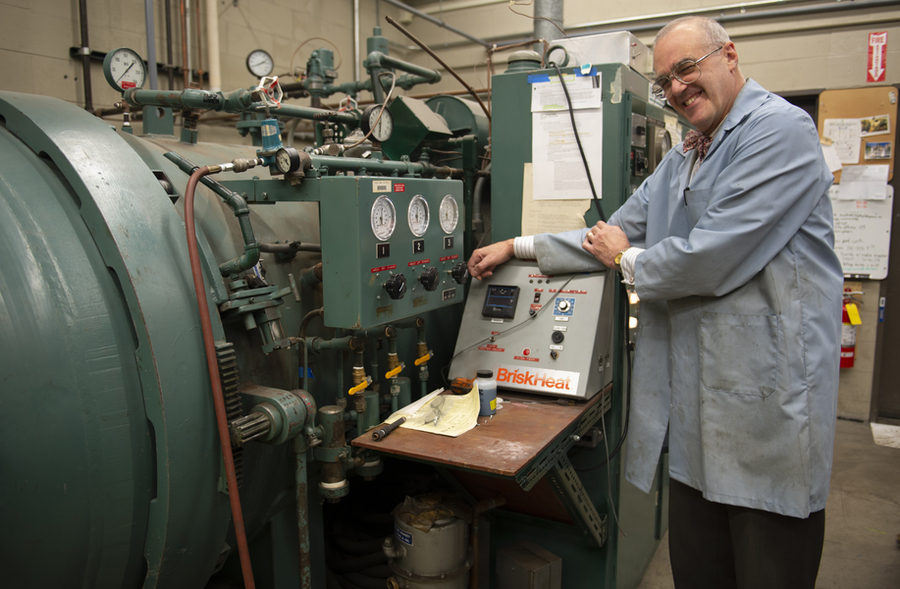 Smiling man in blue lab coat stands in front of large piece of equipment with wires and controls in a lab