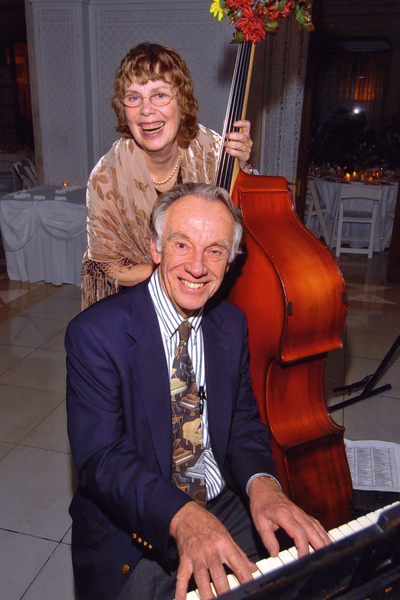 Jake Kerwin at the piano while his wife Marilyn stands behind him, holding her bass. Both are smiling broadly