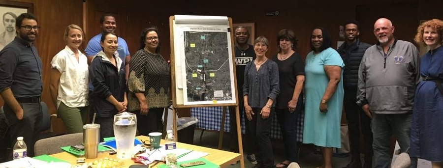 Photo of 12 people standing in a room with a table on the left and an easel with a graphic on it in the center