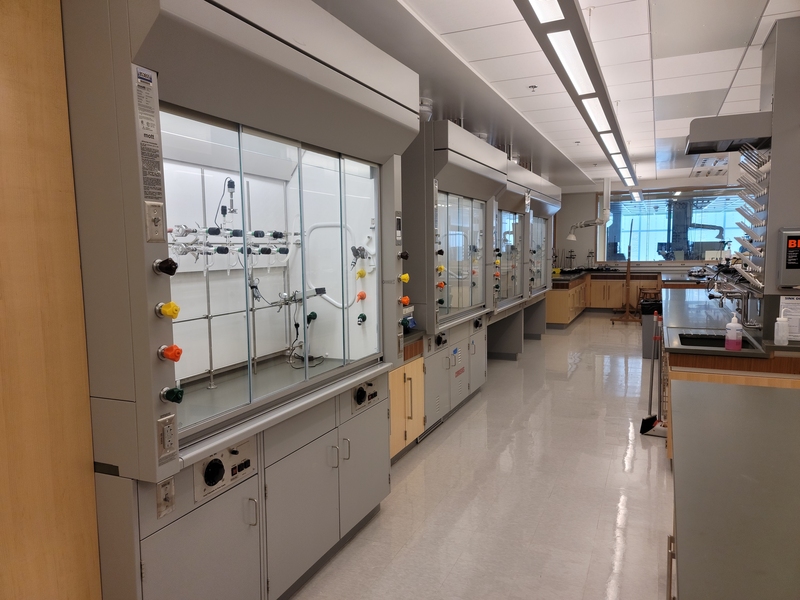 Photo showing some fume hoods in a lab. The hoods are enclosed work stations, several feet across and several feet wide, with pipes and other equipment inside of a glass window