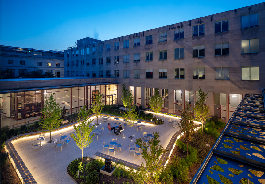 Photo of the Building 14 courtyard as seen from a rooftop at dusk with trees, a curving walkway, and illuminated benches. There are about a dozen circular tables with chairs and two people are seated at one of them.