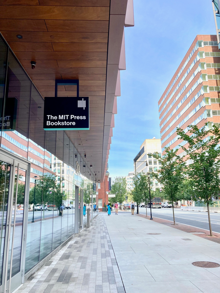 Photo of a side view of a glass-covered building, at left, with a black sign with "The MIT Press Bookstore" hanging from an overhang. The photo looks west with other buildings across the street and short trees along the sidewalk.