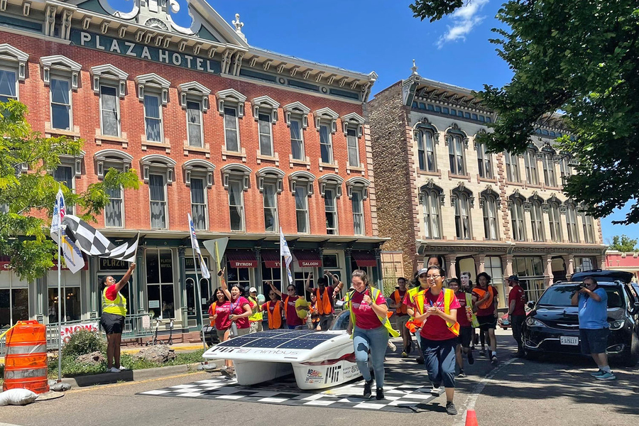 Over a dozen students with crimson t-shirts run, surrounding a solar car, with their hands in the air as they approach a checkered finish line in front of two brick buildings