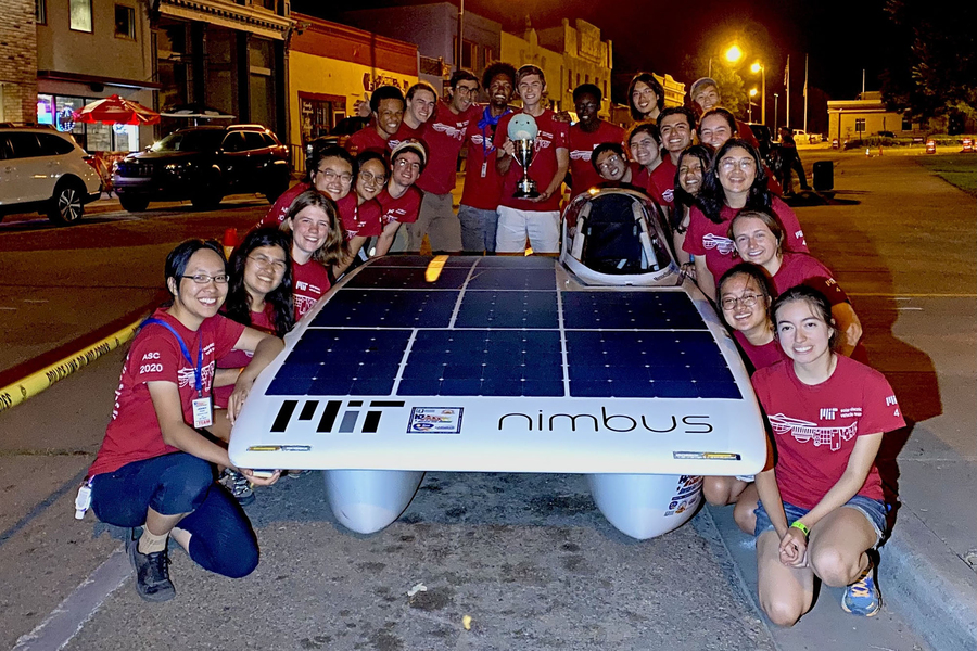Twenty-three students in crimson t-shirts surround a solar car at night, while holding up a trophy