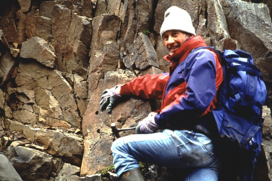 Photo of Fred Frey holding a pick axe and wearing hiking gear while standing next to a rugged rock face