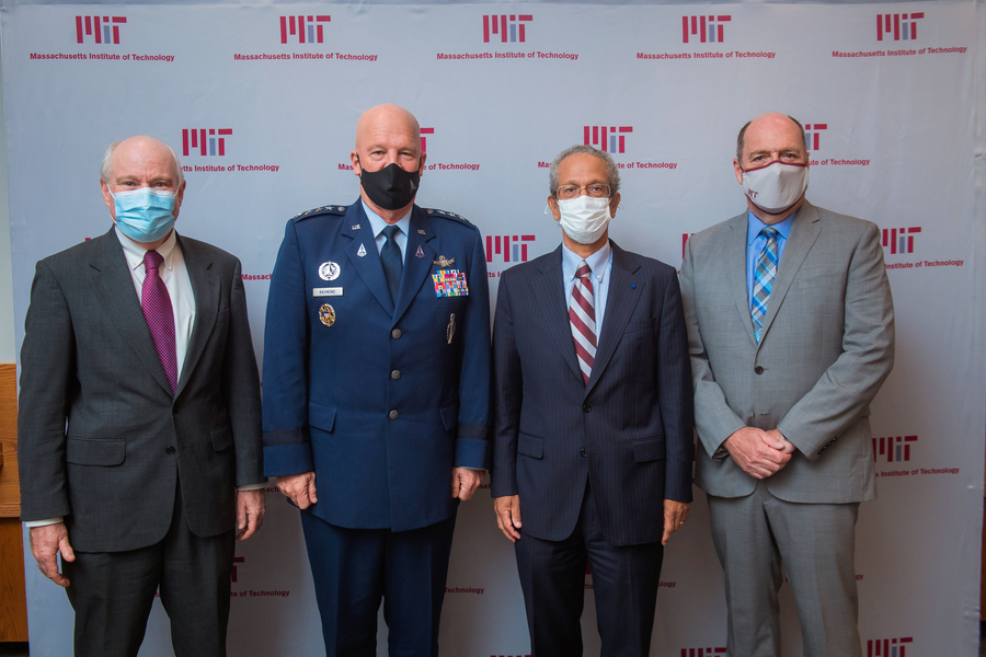 Photo of four men in suits and ties standing together, each wearing a facemask, against a wall printed with repeating "MIT - Massachusetts Institute of Technology"