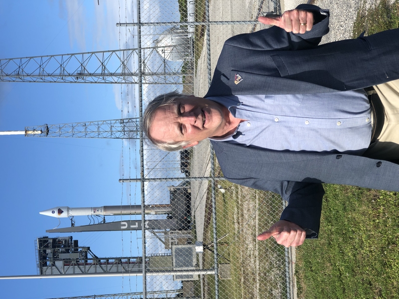 Photo of a man standing in front of a rocket launch site, smiling with two thumbs up