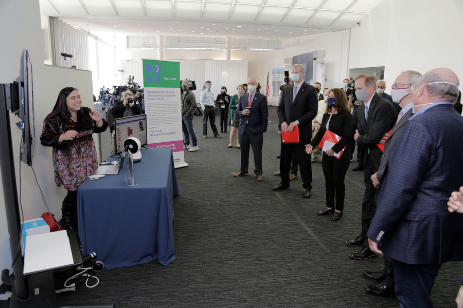 Photo of six people in suits and face masks watching a presentation by a young woman, who is laughing and who is demonstrating with a small spherical robot