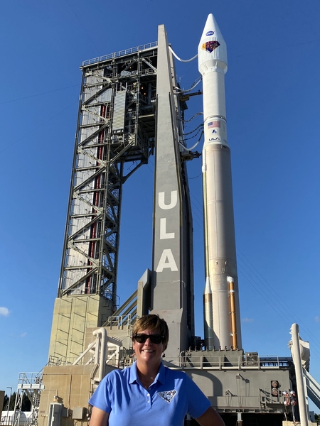 A smiling woman stands immediately in front of a huge rocket launch setup on a sunny day with no clouds