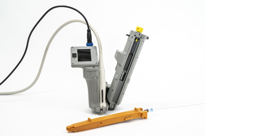 An image of the handheld AI-Guide tool. It consists of a grey plastic device, with a small screen and a needle cartridge; a white ultrasound probe; and an orange application-specific guidewire cartridge.