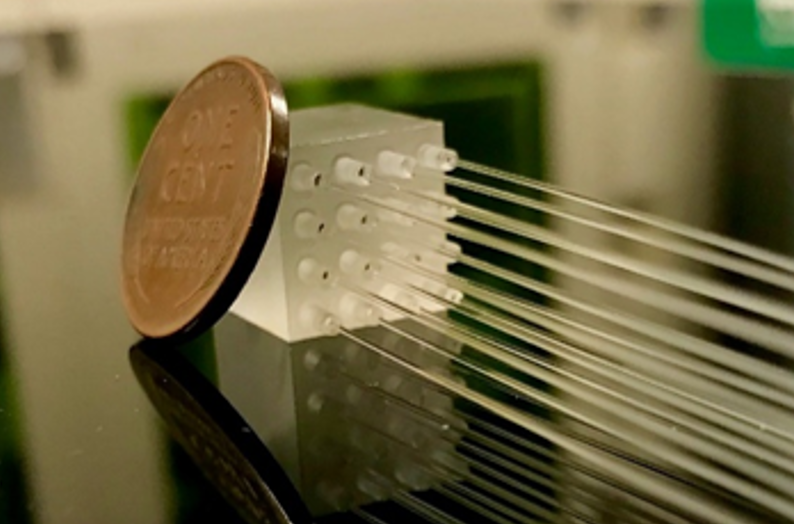 A photo of a small clear glass cube with 16 holes in it, which fiber optics are coming out of. A penny rests against the cube to show its scale.