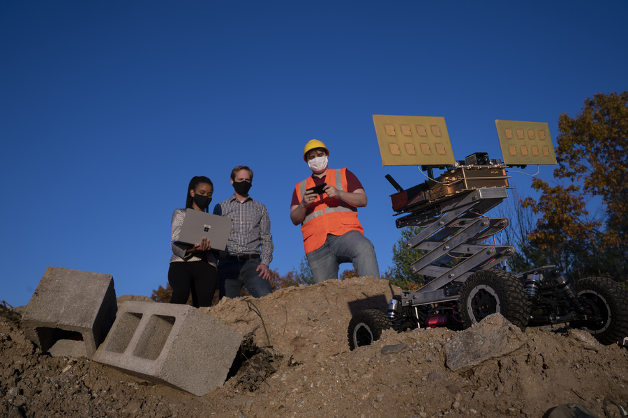 Photo of three people standing on a pile of dirt and construction debris on a sunny day. One researcher is holding a laptop, which another looks at. The third person is holding a small handheld device, and a four-wheeled robotic vehicle is also on the dirt pile.