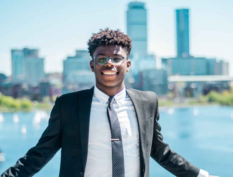 Photo of Dolapo Adedokun, wearing a suit and tie, posing in front of the Charles River on a sunny summer day.