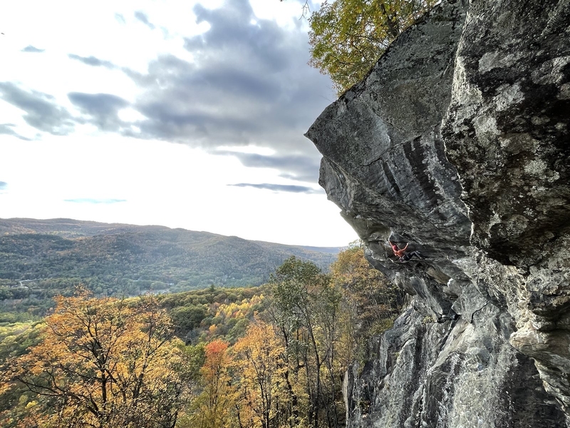 Photo of a person rock climbing a cliff with views of mountains and fall foliage.