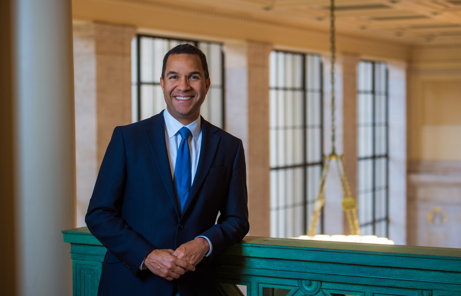 Photo of John Dozier in a suit and tie, smiling and leaning on a railing