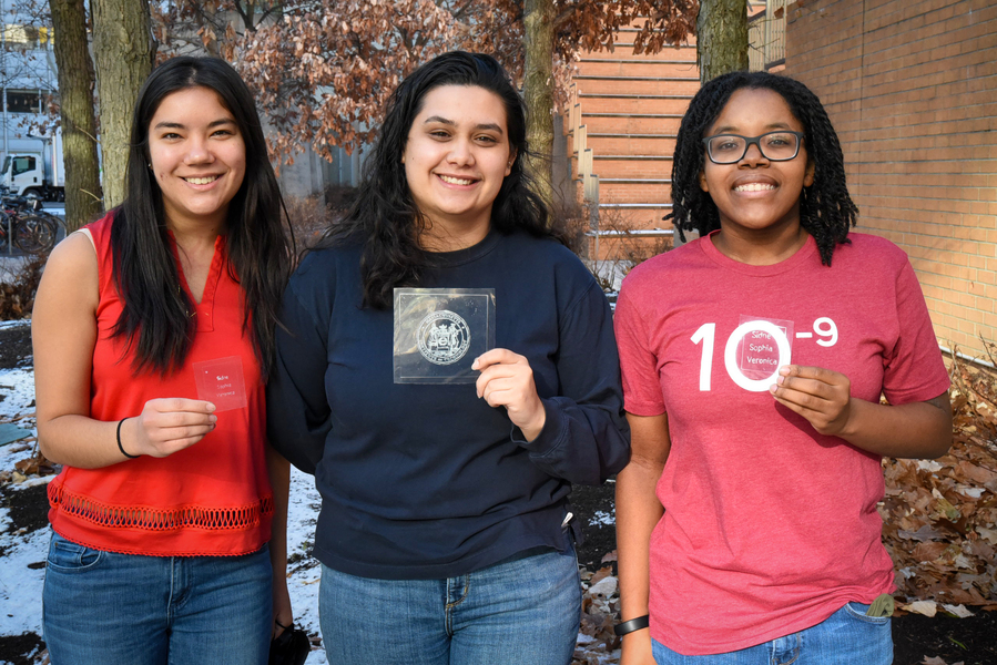 Photo of three female students standing outdoors, holding pieces of imprinted clear plastic