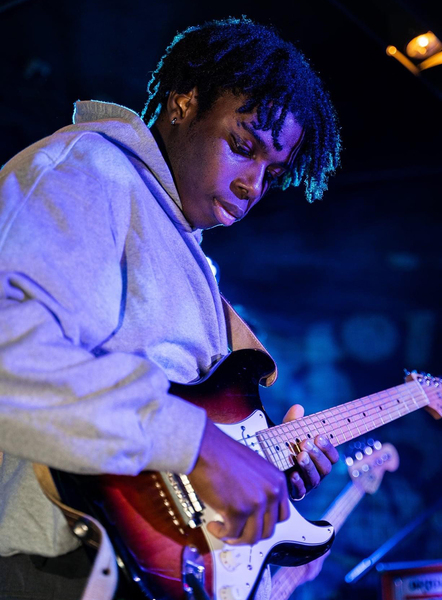 Photo of Dolapo Adedokun with a look of intense concentration on his face as he plays an electric guitar. He is bathed in blue light. 