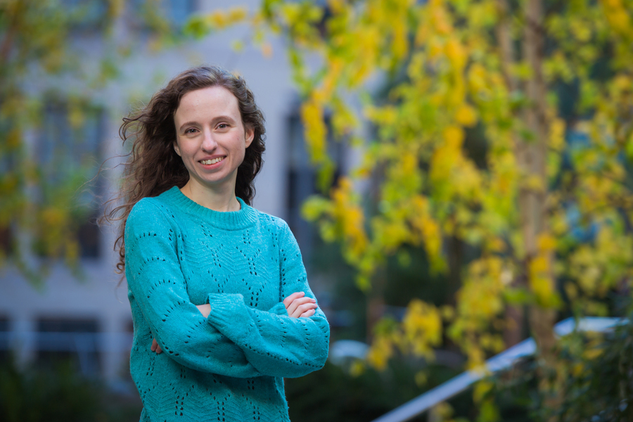 Photo of Nina Andrejević with her arms crossed and a yellow-leafed tree and MIT building in the background