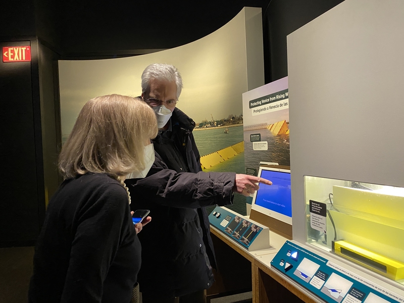 Photo of a man with short grey hair and a mask pointing at a science exhibit that is made of a box filled with water and a yellow barrier resting at the bottom. He is talking to a woman with shoulder-length grey hair who is seen in profile, also looking at the exhibit.