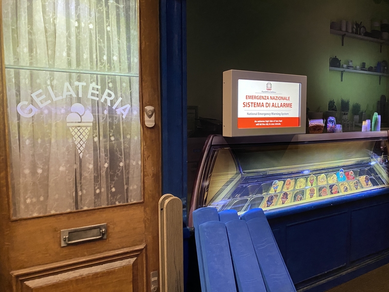 Photo of a mock gelato shop. The door has a glass window with the word “Gelateria” written on it, along with an ice cream cone. Next to the door is a digital screen with red print in Italian, indicating a national emergency.