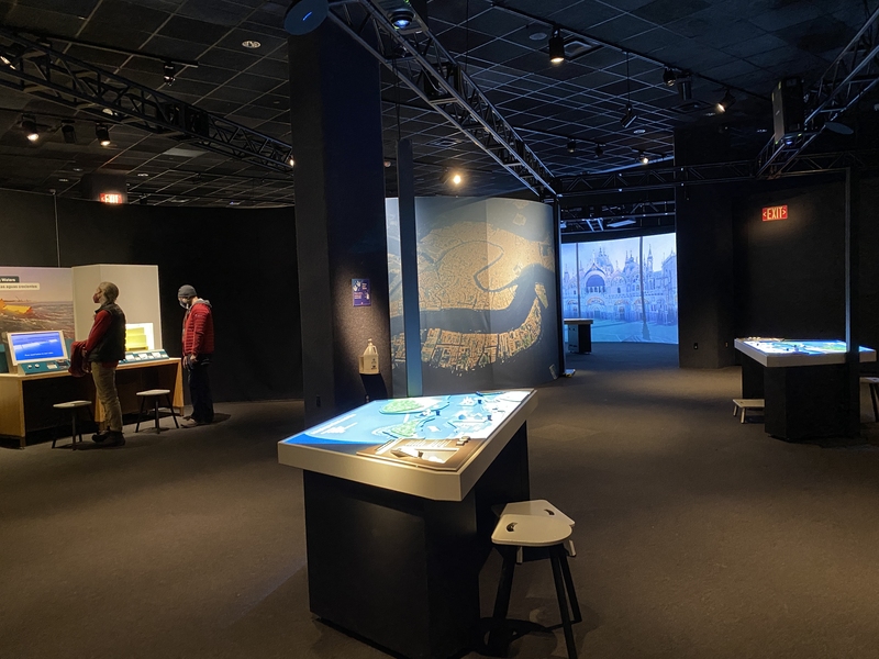 Photo of a large, dark room with high ceilings. In the center of the room is a table with a green and blue projection in it. Behind the table is a large image of the Venetian Lagoon, taken from above. To the left are two people looking at a display with a screen and a box filled with water.