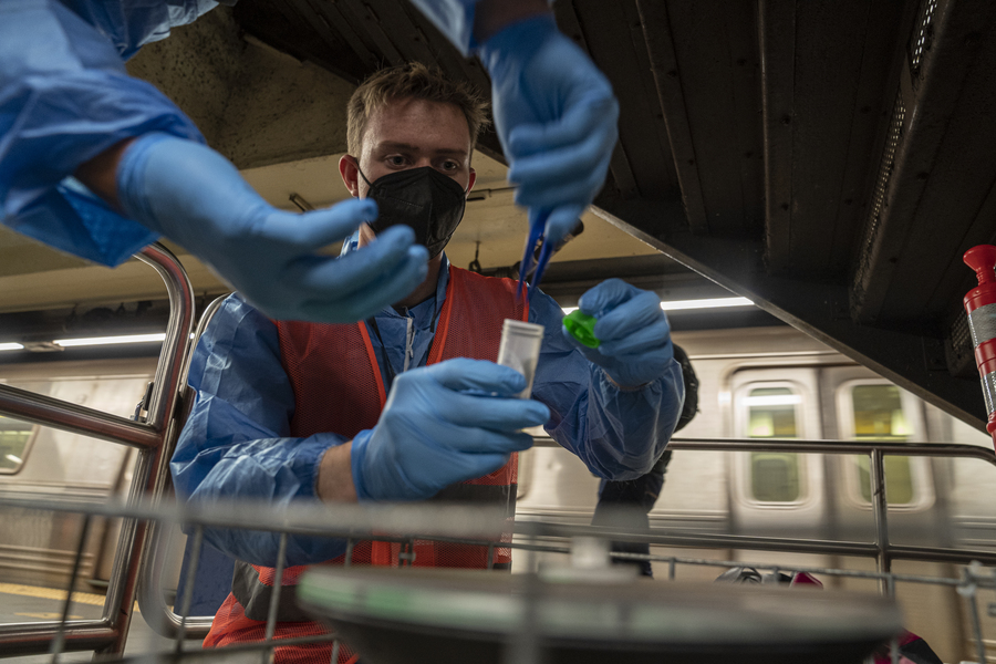 Photo of a researcher in an orange vest and blue gloves putting a small filter into a collection tube (they are helped by their colleague, out of frame except for their hands). They are in a subway station, with a train in view behind them.