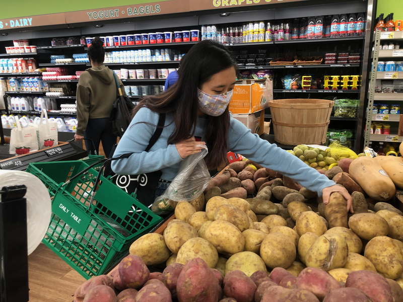 Photo of a masked student holding on to a Daily Table grocery basket and reaching for a potato.