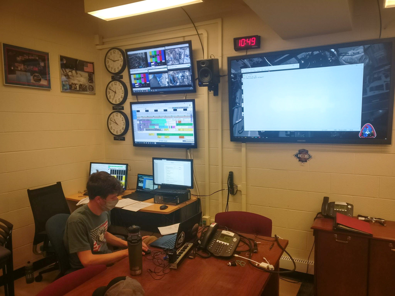 Photo of a masked student looking at computer in front of wall of screens with analog clocks from three time zones