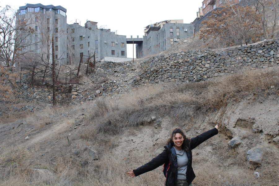 Photo of Zoe Fisher with open arms in front of a hillside topped by a large building