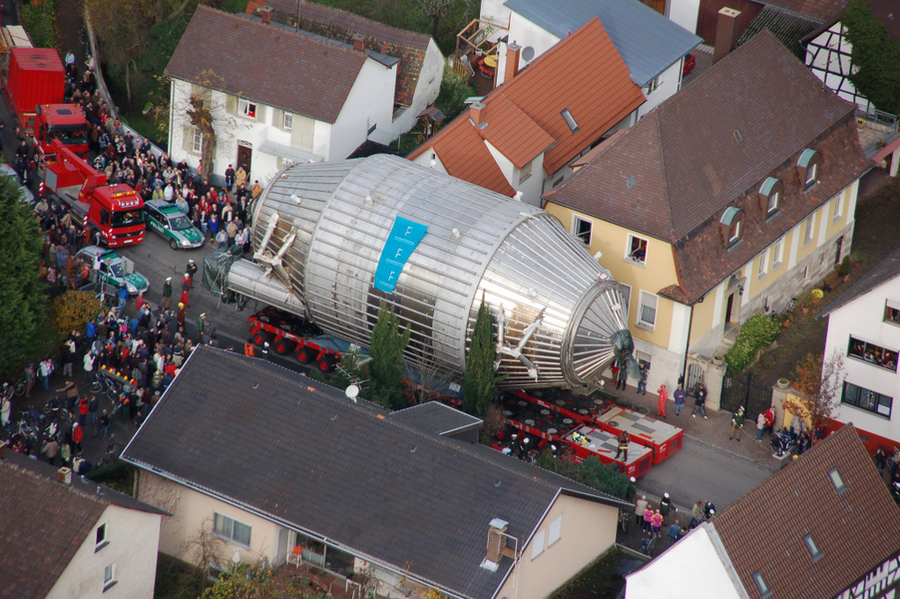 A large metal vessel being transported through a town street; a size comparable to a large 2-story building. A large crowd is gathered to watch.